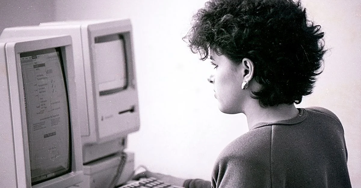 Una mujer usando un ordenador vintage en un entorno de oficina, capturada en escala de grises.