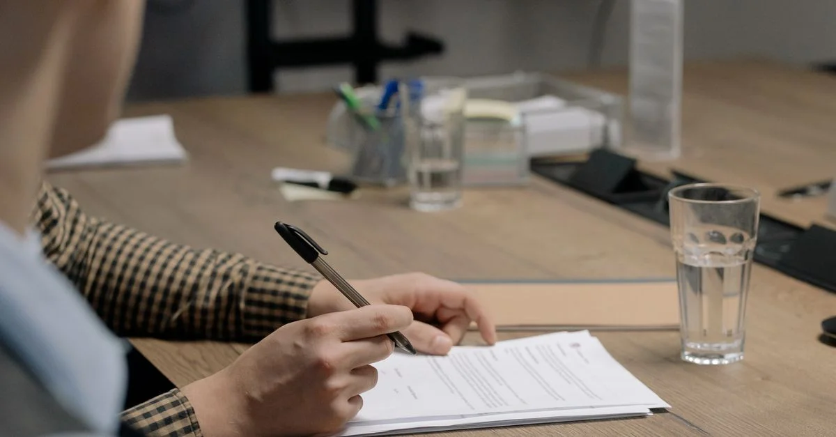 Persona con atuendo de negocios firmando un documento en una mesa de madera en un entorno de oficina.